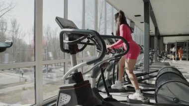 Bir kadın, geniş pencereli, güzel ve modern bir spor salonunda, eliptik bir makine üzerinde gayretle egzersiz yapıyor.