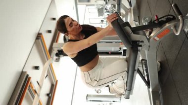 Fitness kızı her egzersiz sırasında formda ve teknikte kalmak için simülatörlerle antrenman yapar. Dikey video.