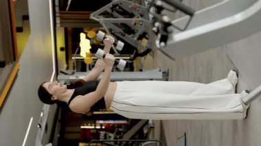 Bir kadın modern ve çağdaş fitness ortamında çeşitli dambıl egzersizleri yapmakla meşgul. Dikey video