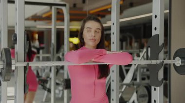 Pembe atletizm kıyafeti giymiş bir kadın şık, modern bir spor salonunda halterle rahatça poz verir..