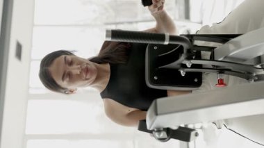 Genç bir kadın, fitness hedeflerine ulaşmak ve sağlığını iyileştirmek için gelişmiş spor aletleri kullanan egzersizlere odaklanıyor. Dikey video