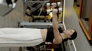 Güçlü bir kadın, modern bir spor salonunda dambıllarla egzersiz yapar. Gücünü ve genel olarak formda olduğunu gösterir.