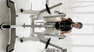 Genç bir kadın simülatör üzerinde gayretli bir şekilde çalışıyor, yoğun bir şekilde güç eğitimine odaklanıyor. Dikey video.