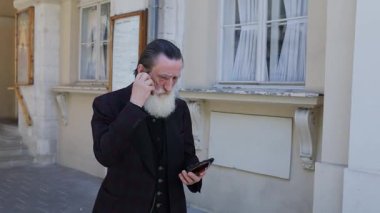 Resmi giyinmiş yaşlı bir adam, açık havada akıllı telefonuyla sohbet ediyor.