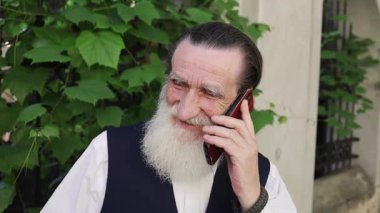 Yaşlı adam canlı yeşil yapraklı arka planda mutlu bir şekilde akıllı telefondan konuşuyor.. 