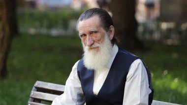 Parktaki bankta oturan uzun beyaz sakallı yaşlı adam. Güneşli açık havada düşünceli bir ifade takınıyor.. 