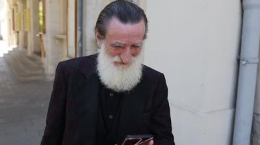 Senior gentleman with long flowing beard thoughtfully uses smartphone in busy urban environment. 