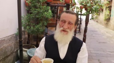 Yaşlı bir adam, yeşilliklerle çevrili rahat bir açık hava ortamında kahvesini yudumluyor..