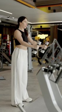 Bir kadın modern ve çağdaş fitness ortamında çeşitli dambıl egzersizleri yapmakla meşgul. Dikey video