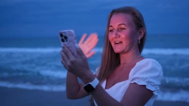 Alacakaranlık sırasında kumsalda telefonuyla neşeli bir şekilde meşgul olan bir kadın modern iletişimi sergiliyor..