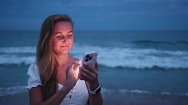Bir kız alacakaranlıkta sakin deniz kenarında akıllı telefonunu neşeyle kullanır, ekranda hafifçe aydınlatılır..
