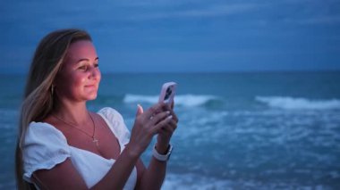 Neşeli bir gülümsemesi olan bir kadın, büyüleyici alacakaranlık saatinde okyanusun kenarında dururken akıllı telefonunu kullanır.