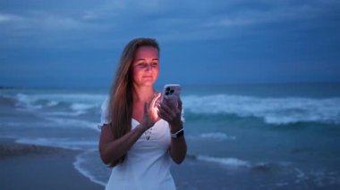 Kumsalda dalgalı uzun saçlı bir kadın dalgalar arkasındayken akıllı telefonunu kullanıyor.