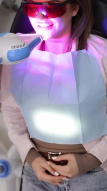 Güzel bir gülümseme geliştirmek için LED teknolojisi kullanan bir kadın diş beyazlatma prosedüründen geçiyor. Dikey video.