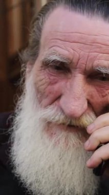 Uzun sakallı yaşlı bir adam açık havada telefonla konuşuyor. Dikey video.