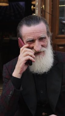 Kentin dışında güneşli bir günün tadını çıkarırken akıllı telefondan konuşan kendinden emin yaşlı sakallı bir adam. Dikey video.