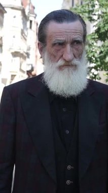Beyaz sakallı, kendine güveni tam yaşlı bir adam. Büyüleyici klasik caddelerde geziniyor. Dikey video. 