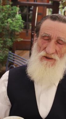 Yaşlı bir adam yemyeşil yeşilliklerle dolu rahat bir ortamda kahve içiyor. Dikey video. 