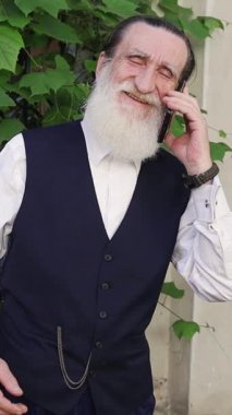 Geleneksel giyinmiş yaşlı adam yeşil bahçede telefonla konuşuyor. Dikey video. 