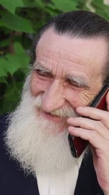 Yaşlı adam yeşil yapraklarla çevrili akıllı telefondan mutlu bir şekilde konuşuyor. Dikey video. 
