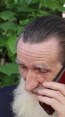 Etrafı yemyeşil bitkilerle çevrili, uzun beyaz sakallı yaşlı bir adam telefonda konuşuyor. Dikey video. 