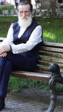 Parktaki bankta oturan uzun beyaz sakallı yaşlı bir adam. Güneşli açık hava ortamında düşünceli bir yüz ifadesi var. Dikey video.