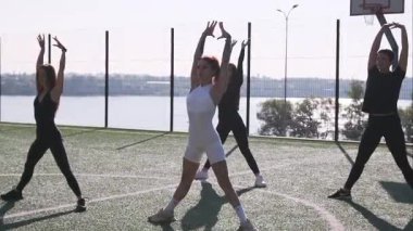 Bir grup kadın güneşin altındaki yeşil çimenlikte dinamik egzersiz hareketleri sergiliyor, dışarıda eğlenceli bir spor salonunda güç, koordinasyon ve pozitif enerji sergiliyor..