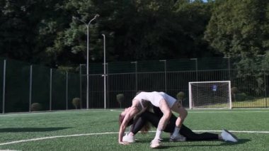 İki atletik kadın güneşli bir futbol sahasında enerjik fitness hareketleri yapıyorlar. Açık hava antrenmanında güç, koordinasyon ve takım çalışması sergiliyorlar..