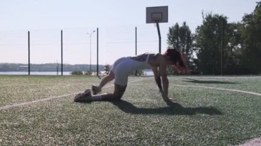 Formda bir kadın, güneşli bir futbol sahasında derin esneme egzersizleri yapar. Denge, esneklik ve sağlığını doğal ortamlarda gösterir..