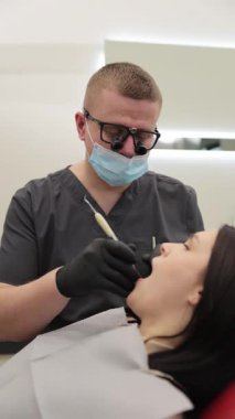 Bir hasta modern bir klinikte gelişmiş ekipman kullanan çok yetenekli ve profesyonel bir dişçiden kapsamlı bir diş muayenesi alıyor. Dikey video.