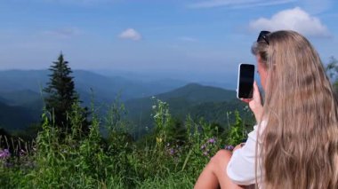 Genç bir kadın akıllı telefon kamerasıyla nefes kesici bir dağ manzarası yakalıyor.