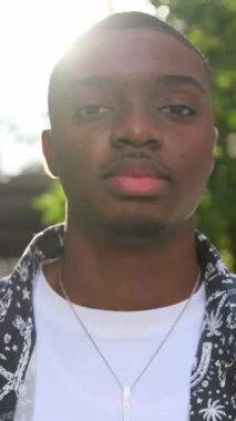 Yeşil ve güneş ışığıyla çevrili afro-amerikan adam kendine güvenen bir şekilde dışarıda duruyor. Dikey video.