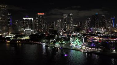 Miami siluetinin gece hava aracı görüntüleri. Dönme dolap, gökdelenler ve körfezde parlayan yansımalar var.