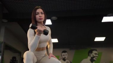 Seksi genç esmer, bir eliyle spor salonunda dambılı kaldırıyor. Genç bir kadının aktif eğitimi. Güzel bir vücudun ve figürün konseri.