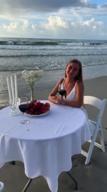 Okyanus kıyısında beyaz bir masada oturan ve şarap içen güzel genç bir kadın. Okyanus kıyısında güzel bir yer