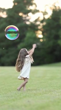 Beyaz elbiseli genç bir kız çimenli bir tarlada oynarken bir sabun köpüğünü yakalamak için uzanıyor. Dikey video