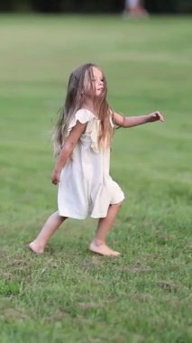 Beyaz elbiseli küçük bir kız enerji ve neşe dolu yeşil bir alanda koşuyor. Çocuklar güneşli bir günde dışarıda oynuyorlar..