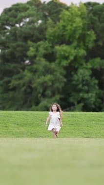 Beyaz elbiseli küçük bir kız doğada geniş bir çim tarlasında neşeyle koşuyor. Dikey video