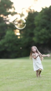 Beyaz elbiseli küçük bir kız neşeyle atlıyor ve çimen tarlasında büyük bir baloncuğa uzanıyor. Dikey video.