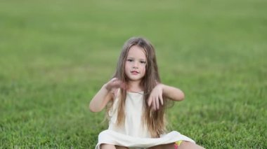Beyaz elbiseli neşeli bir genç kız çimlerin üzerinde oturuyor ve kolları neşeyle kalkıyor. Tatlı bir çocukluk anı..