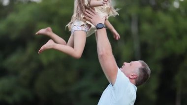 Gülümseyen bir baba açık havada güneşli bir günde küçük kızını neşeyle havaya kaldırıyor. Aile mutluluğu ve bağlılık.