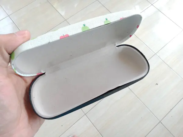Beyaz çiçek desenli kapaklı bir gözlük kılıfı ve beyaz arka planda bir insan eli tarafından tutulan sade siyah bir taban.
