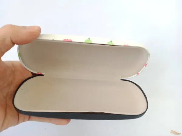 Beyaz çiçek desenli kapaklı bir gözlük kabı ve beyaz arka planda bir insan eli tarafından tutulan sade siyah bir taban.