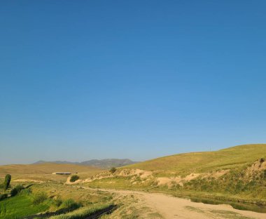 Açık mavi bir gökyüzünün altında yeşil tepeler arasında dolanan toprak bir yolun yer aldığı pitoresk bir kırsal alan. Ağaçlar ve çalılar doğal aksanlar ekler. Yalnızlık ve doğa ile uyum ortamı yaratır..