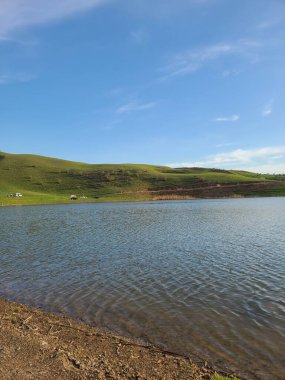 Mavi gökyüzünün altında yuvarlanan yeşil tepeleri olan sakin bir göl.