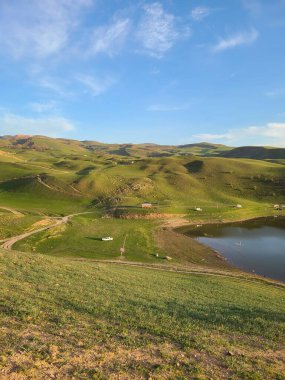 Açık mavi gökyüzünün altında küçük gölü olan manzaralı yeşil tepeler