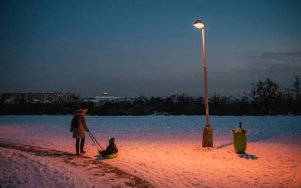Prag 'da bir sokak lambasının altında çocuğunu kızakla çeken genç bir anne vardı. Arka planda prefabrik apartman blokları var. Huysuz fotoğraf.