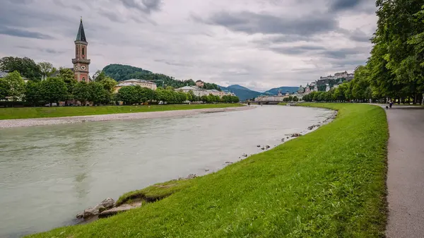 Avusturya 'nın Alp şehri Salzburg' un panoramik manzarası. Nehir boyunca güzel bir gezinti alanı. Tarihi binalar ve arka planda bir kilise..