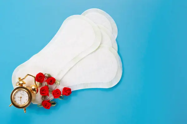 Alarm saati, çiçekler ve mavi arka planda kadın hijyen pedleri. İlk adet dönemi konsepti, adet döngüsü.