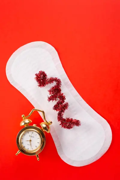 Kırmızı arka planda çalar saat ve kadın hijyen pedi. İlk adet dönemi konsepti, adet döngüsü..